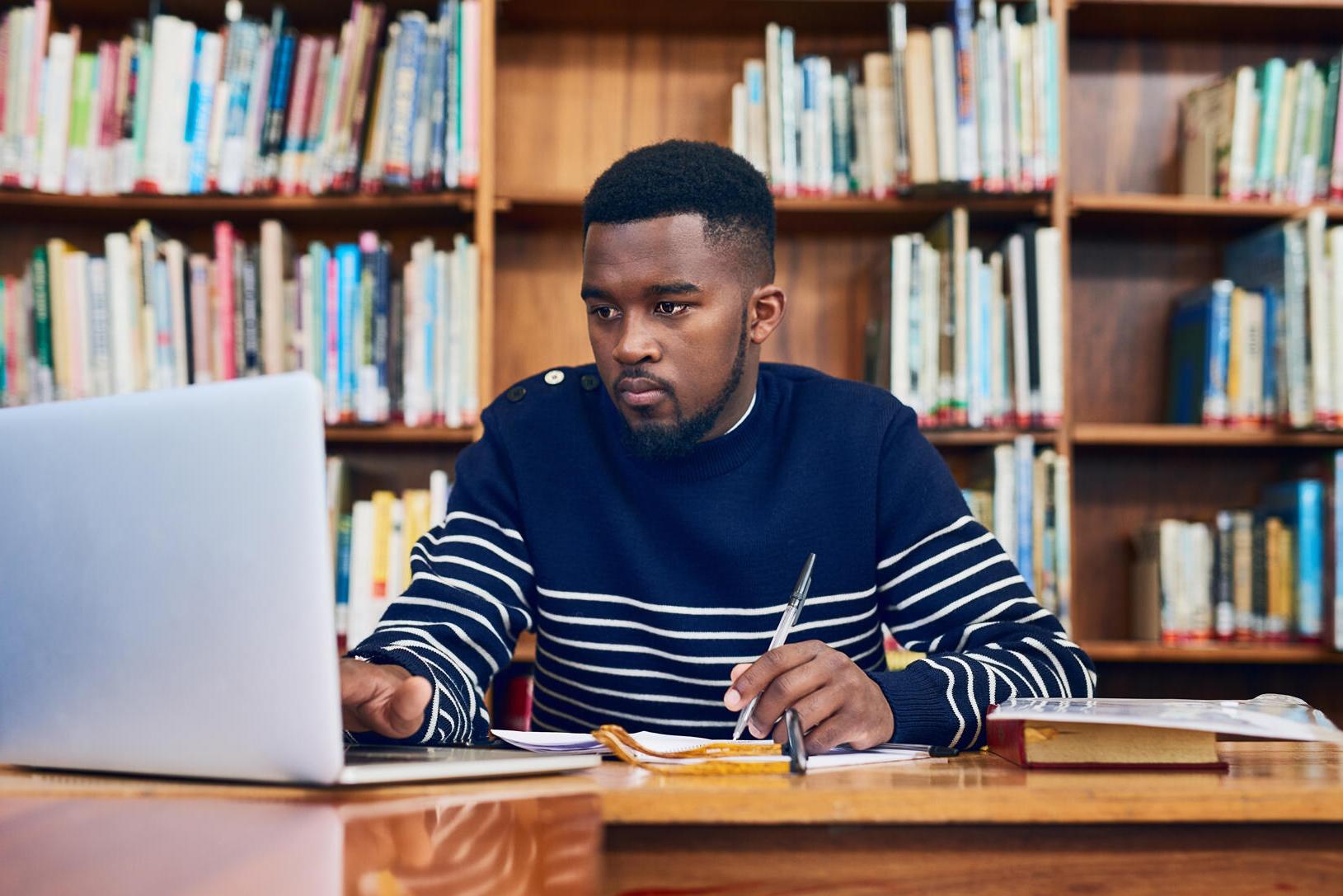 穿着蓝白条纹毛衣的男学生坐在电脑前，后面是书架